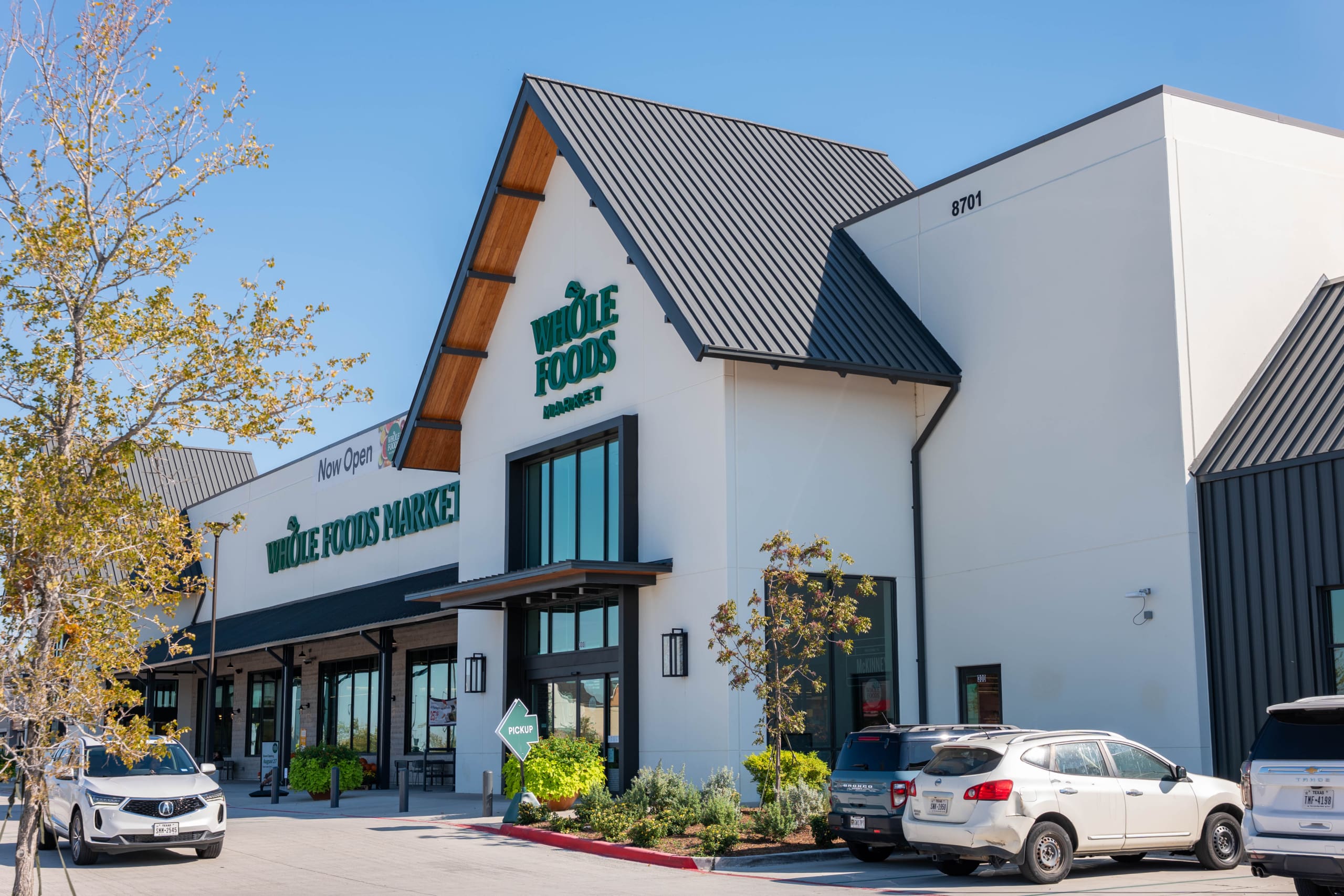 Exterior view of a Whole Foods Market store on a sunny day, with parked cars in front and a "Now Open" sign above the entrance.