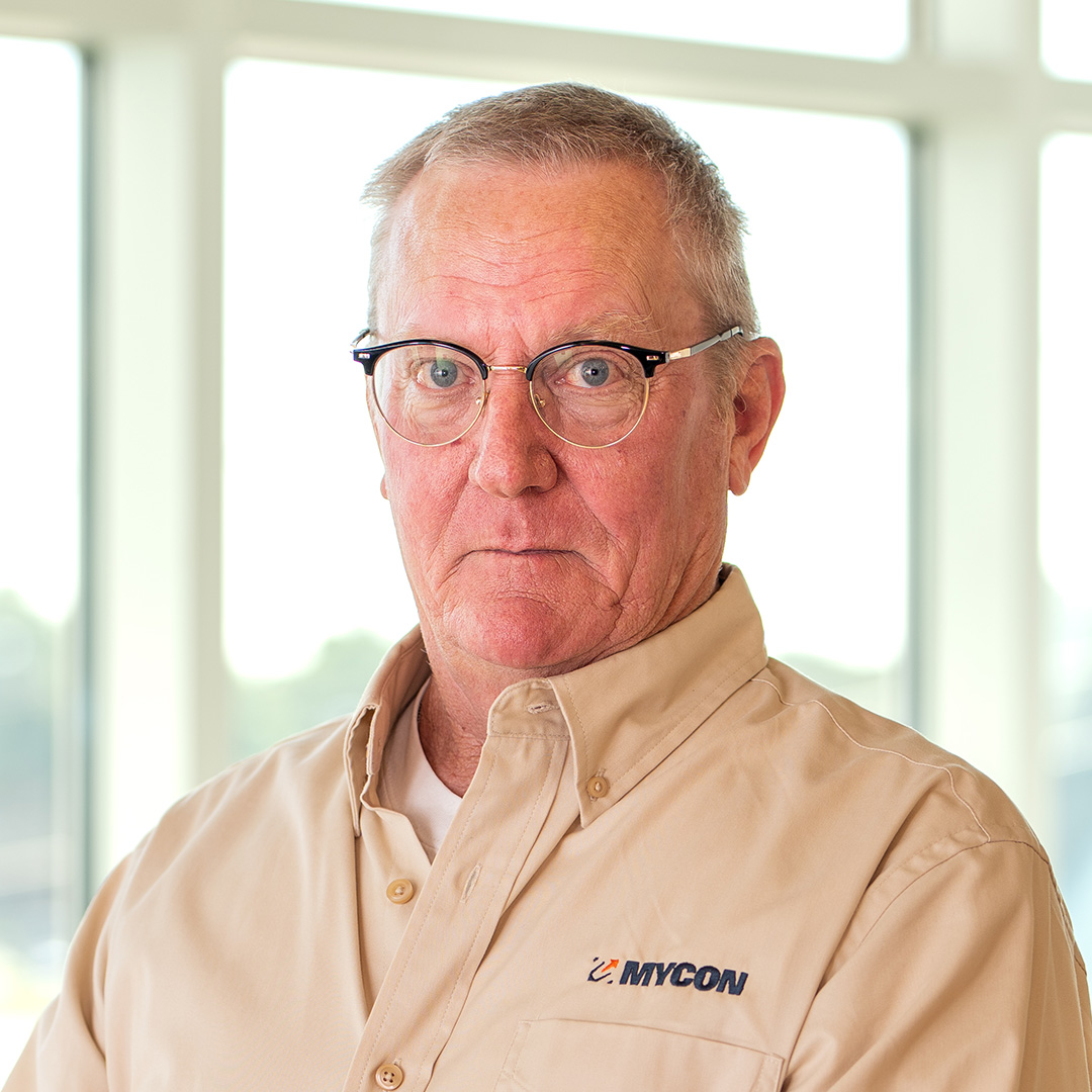 Older man with short gray hair, glasses, and a beige button-up shirt with the "MYCON" logo stands indoors in front of large windows.