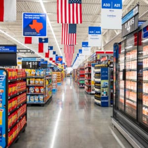 A wide aisle in a brightly lit supermarket near Home Depot San Antonio West Construction displays snacks on shelves and refrigerated drinks, with American and Texas flags hanging from the ceiling.