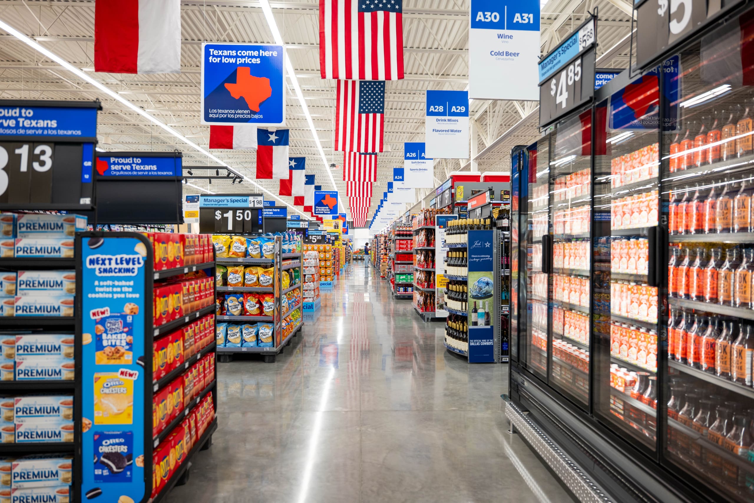 A wide aisle in a brightly lit supermarket near Home Depot San Antonio West Construction displays snacks on shelves and refrigerated drinks, with American and Texas flags hanging from the ceiling.