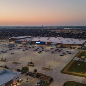 Aerial view of a Walmart store with a large, mostly empty parking lot at sunset, near the Home Depot San Antonio West Construction site, surrounded by suburban houses and roads.