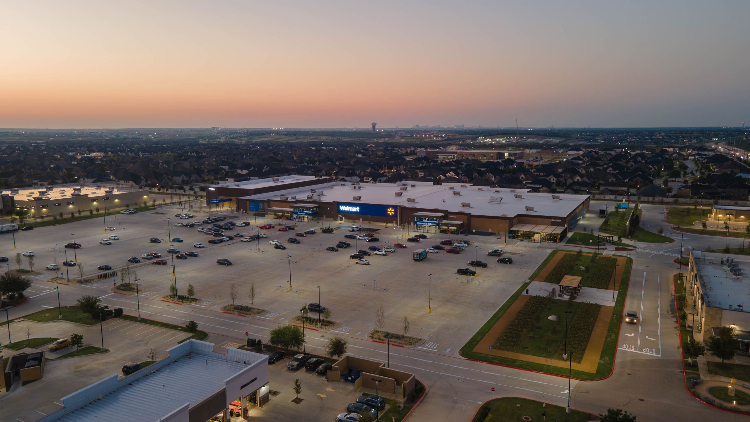 Aerial view of a Walmart store with a large, mostly empty parking lot at sunset, near the Home Depot San Antonio West Construction site, surrounded by suburban houses and roads.