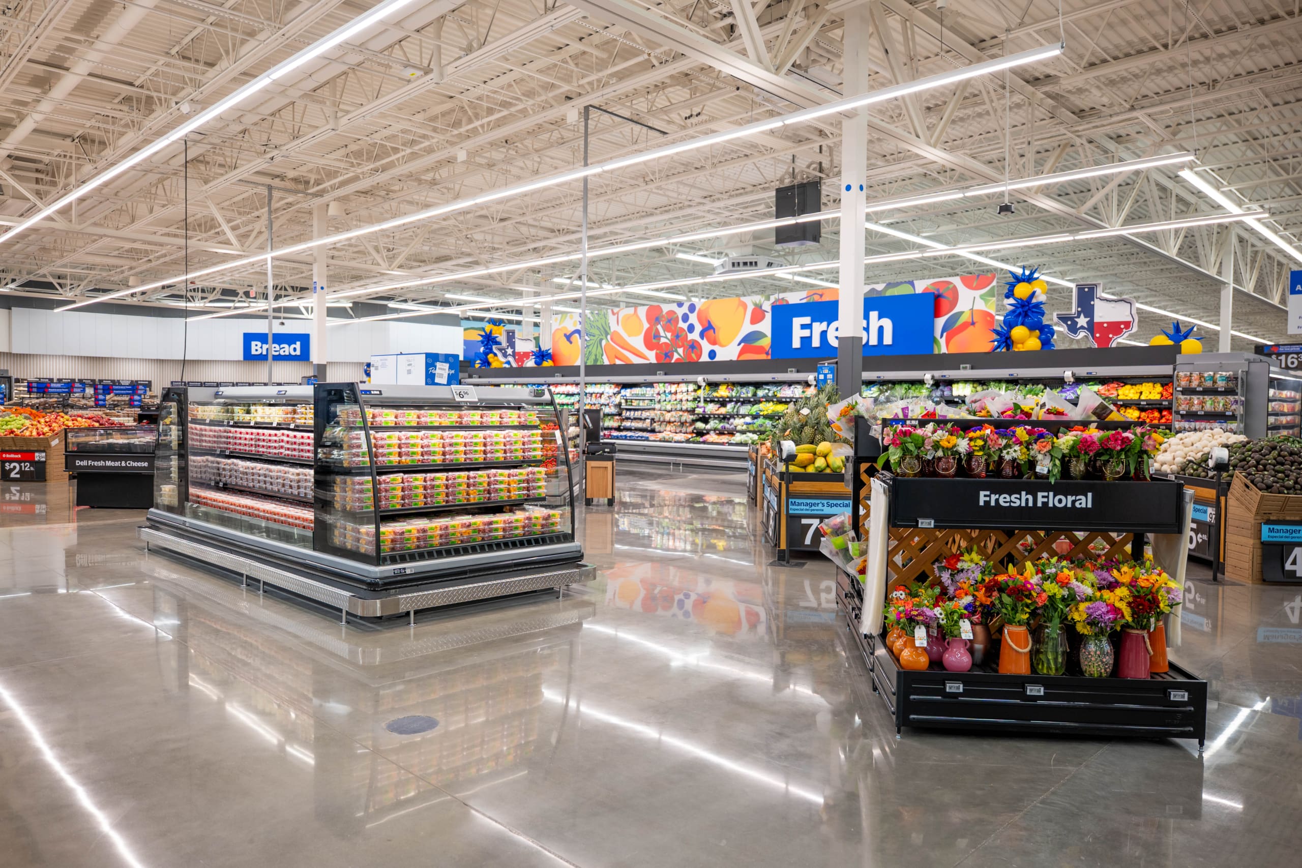 A brightly lit grocery store produce section displays fresh fruits, vegetables, pre-packaged salads, and a stand of assorted floral bouquets, reminiscent of the vibrant atmosphere at Home Depot San Antonio West Construction.