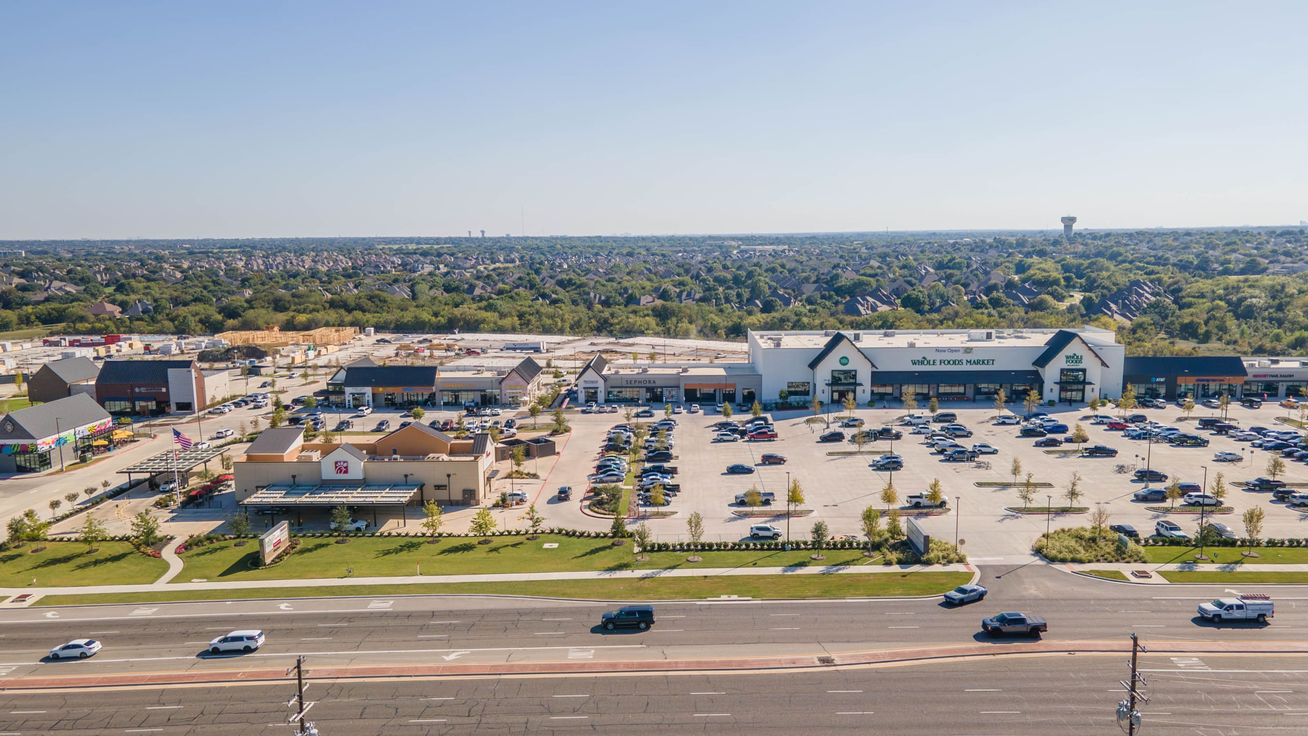 Aerial view of a shopping center featuring a large grocery store, several smaller businesses, and a parking lot, bordered by a busy road and near the site of the Construction, surrounded by suburban neighborhoods.