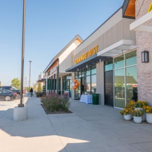 A modern shopping center sidewalk with storefronts, parked cars, flower pots, and balloons near the entrance of a business named “ORANGE TWIST,” on a sunny day.