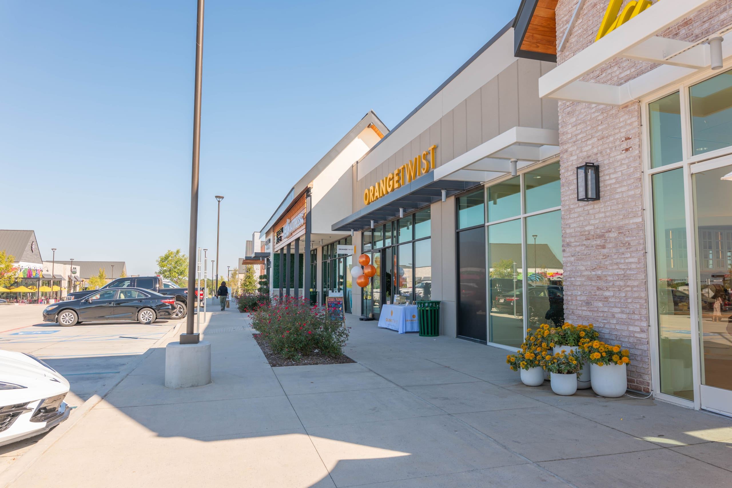 A modern shopping center sidewalk with storefronts, parked cars, flower pots, and balloons near the entrance of a business named “ORANGE TWIST,” on a sunny day.
