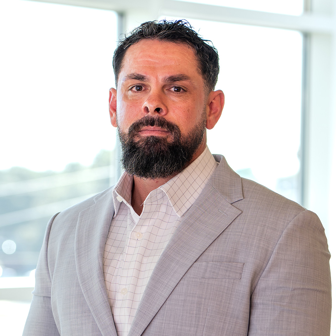 A man with short dark hair and a beard wearing a light gray suit jacket and a white shirt stands indoors with large windows in the background.