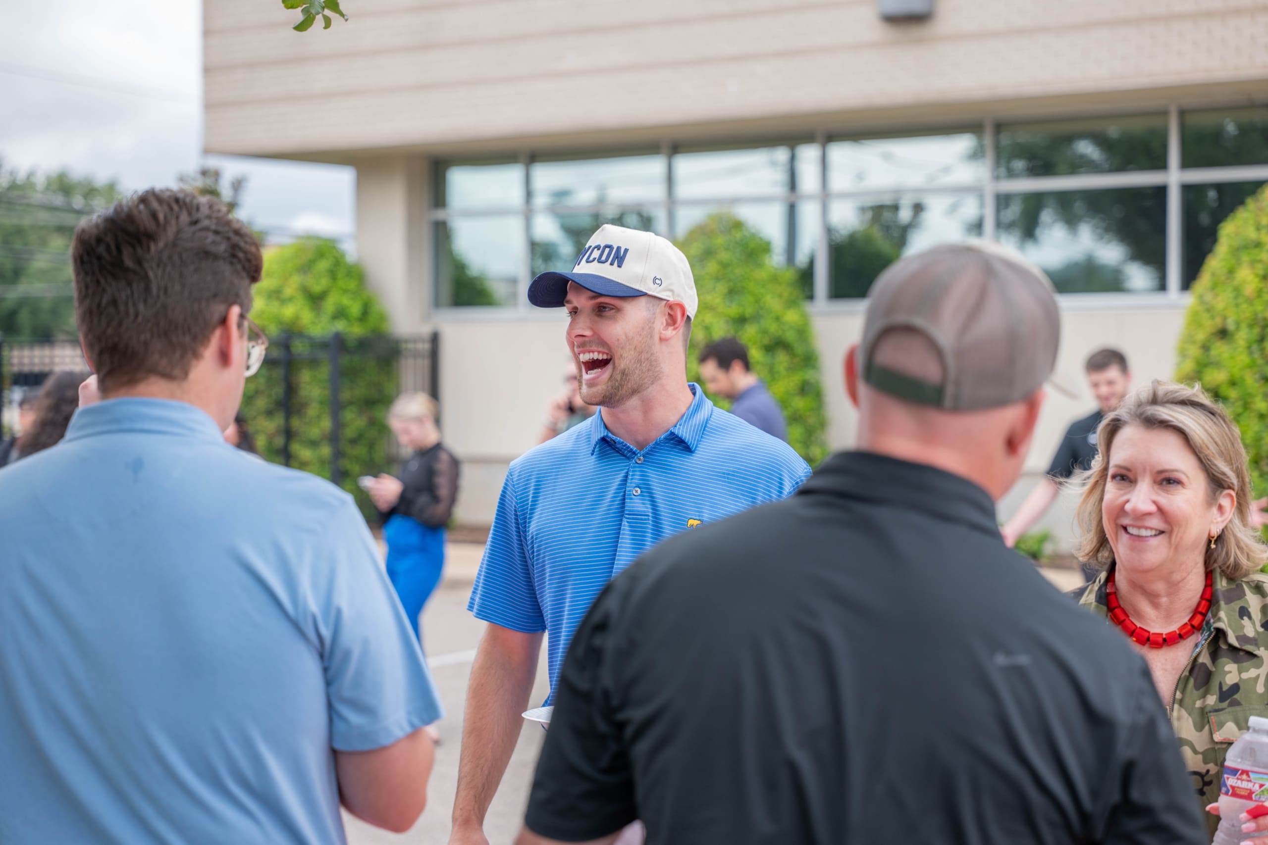 A group of adults converse outdoors near a building, possibly discussing projects with general contractors near me; one man in a white cap and blue shirt is laughing.