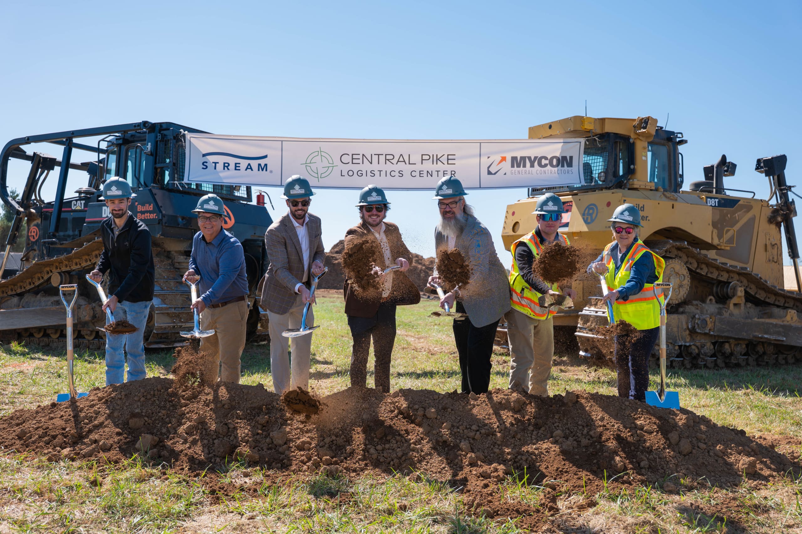 MYCON Employees in construction gear take part in a groundbreaking ceremony, holding shovels, with excavators and a banner for general contractors near me visible in the background.