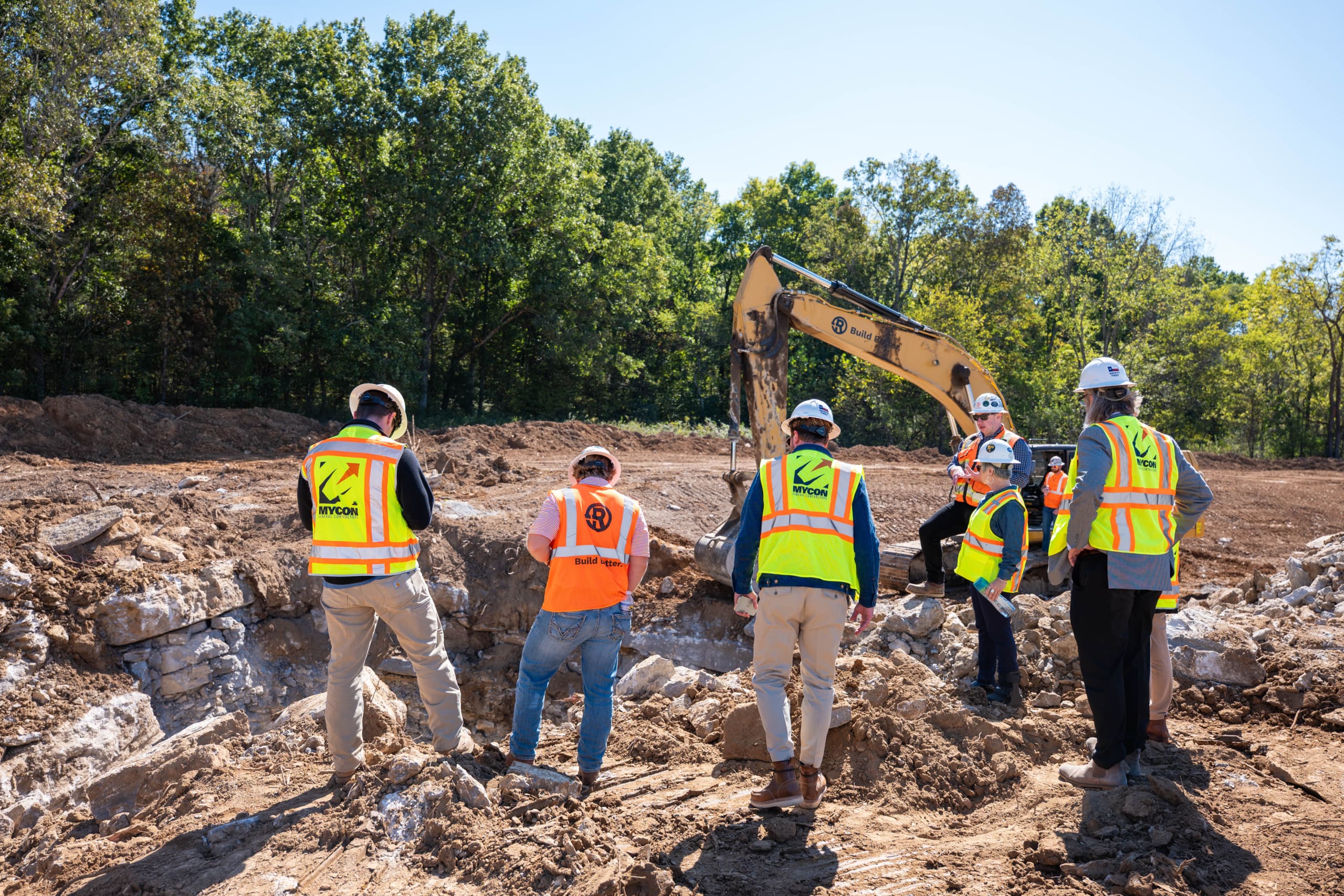 Six construction workers in safety vests and helmets stand at an excavation site, observing a backhoe in operation amid dirt and rocks, with trees in the background—ideal expertise for those seeking reliable general contractors near me.