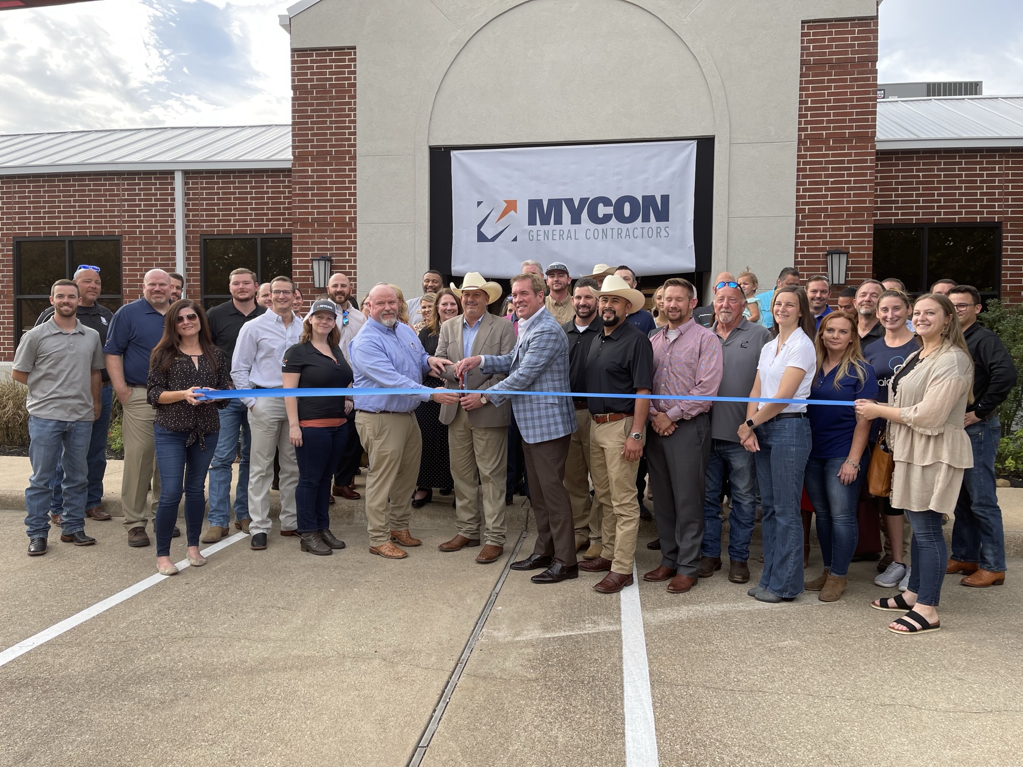 A group of people gathers outside a brick building for a ribbon-cutting ceremony in front of a MYCON General Contractors banner, highlighting trusted general contractors near me.