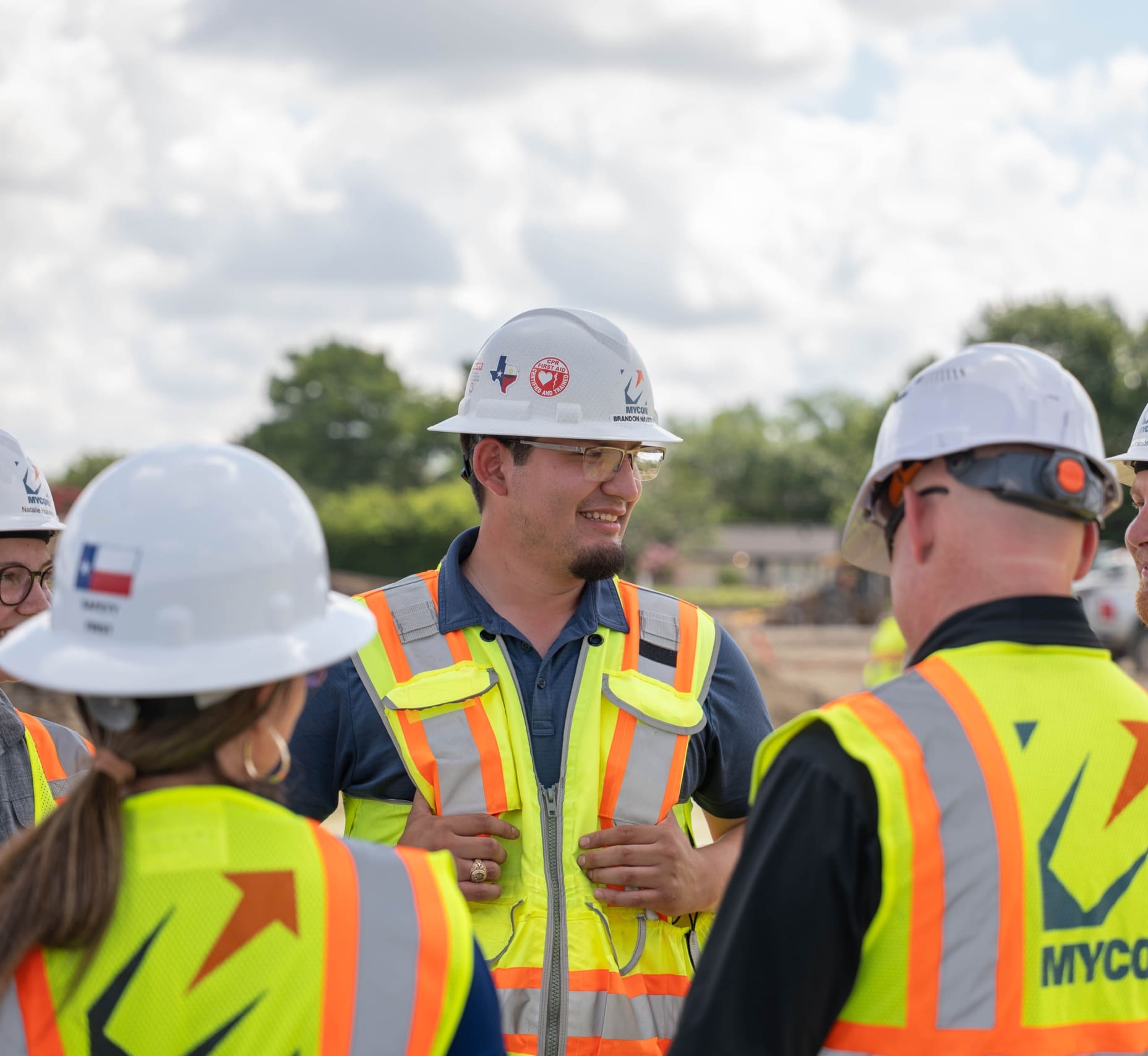 A group of construction workers in safety vests and hard hats stand together outdoors, talking at a work site—typical of teams provided by general contractors near me.