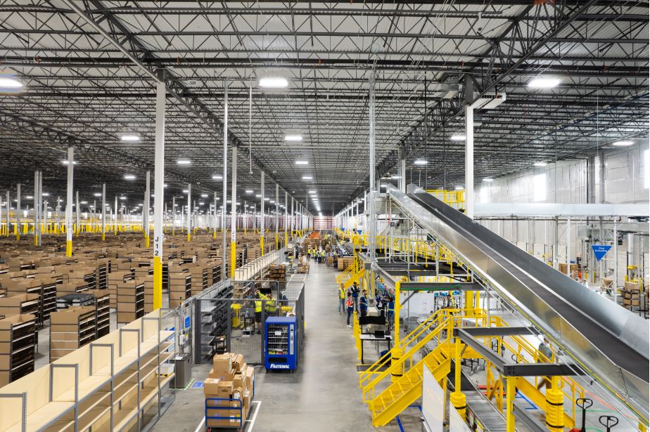 Large warehouse interior showcases Retail Construction Expertise with tall shelves of boxes, conveyor belts, and workers operating among organized packaging and shipping stations under bright lighting.