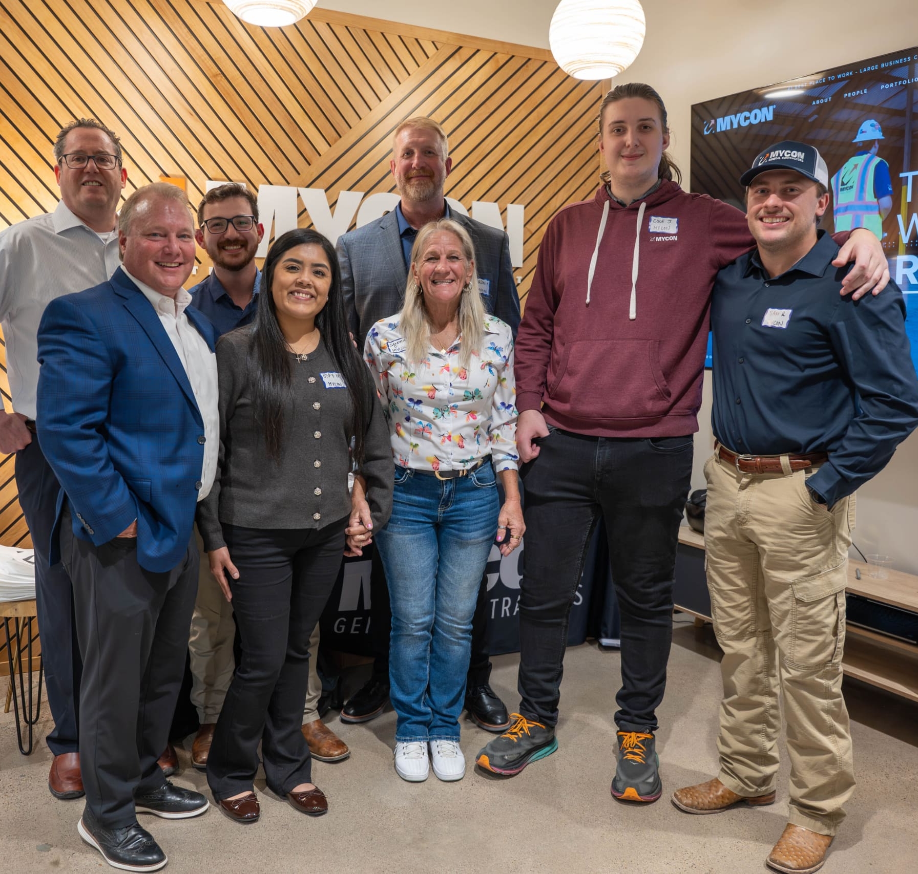 A group of eight people stand together indoors, posing for a photo in front of a MYCON sign and a television displaying construction-related content—perfect for those searching for reputable general contractors near me.