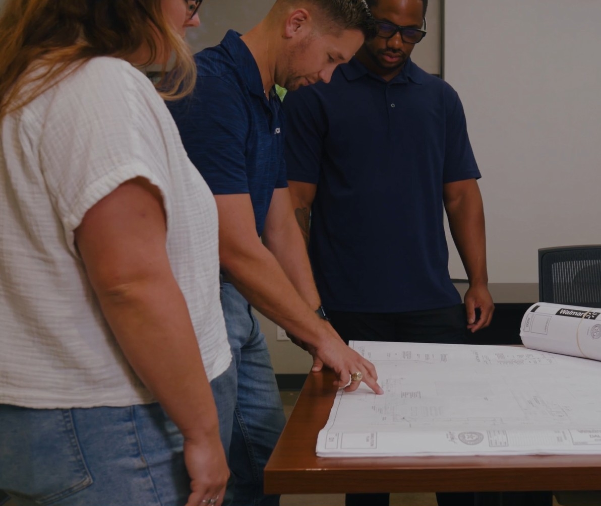 Three people stand around a table, reviewing and pointing at architectural blueprints—an ideal scene for those searching for general contractors near me.