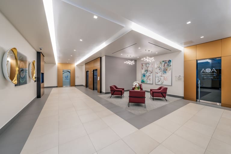 Modern office lobby with three red chairs around a small table, two elevators, wall art, and a glass door labeled "Orthopedic Associates of Houston," located near convenient self storage options.