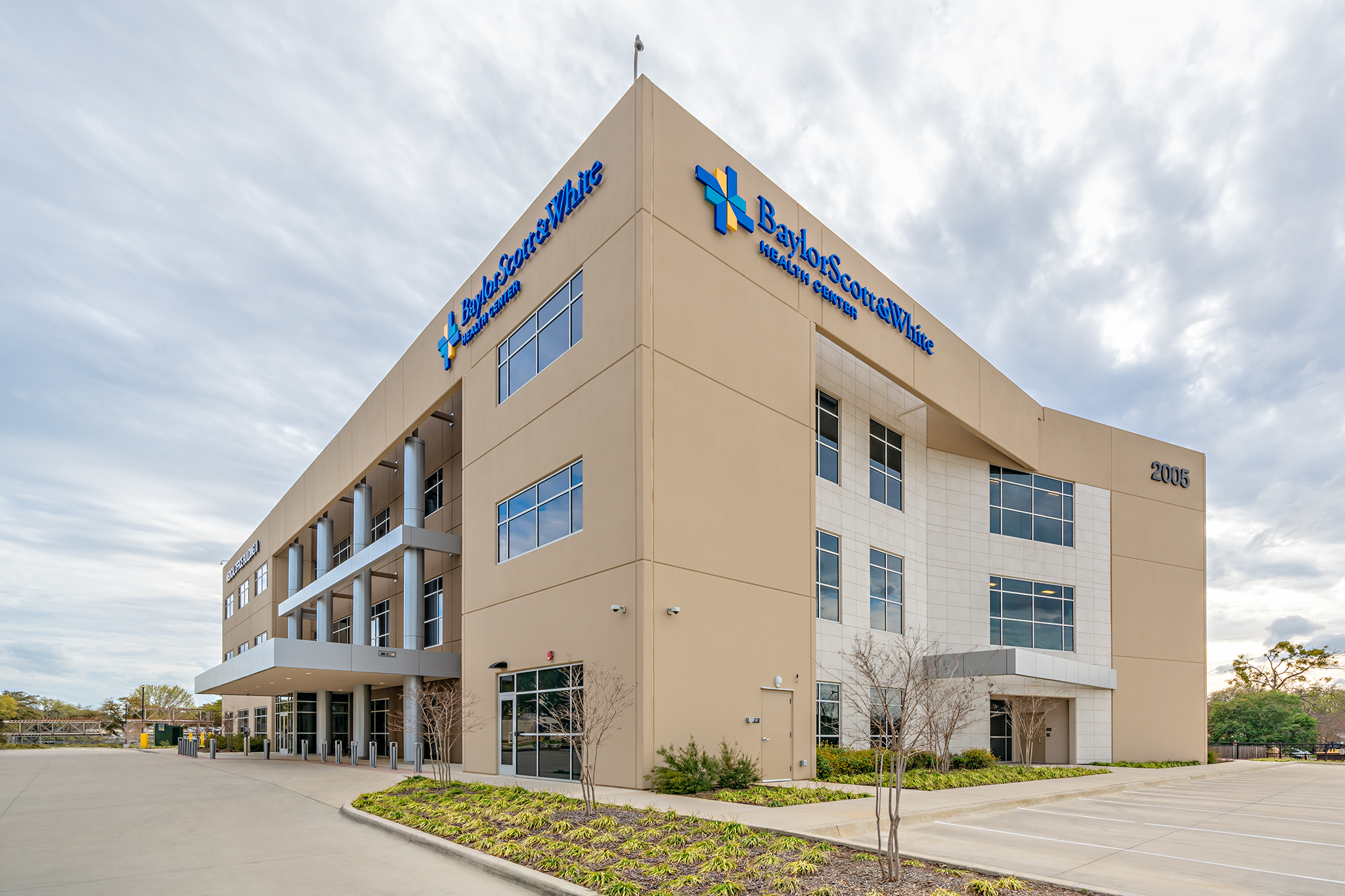 A three-story Baylor Scott & White Health building with large windows, beige and white exterior, and a self storage facility visible near the parking lot under a cloudy sky.