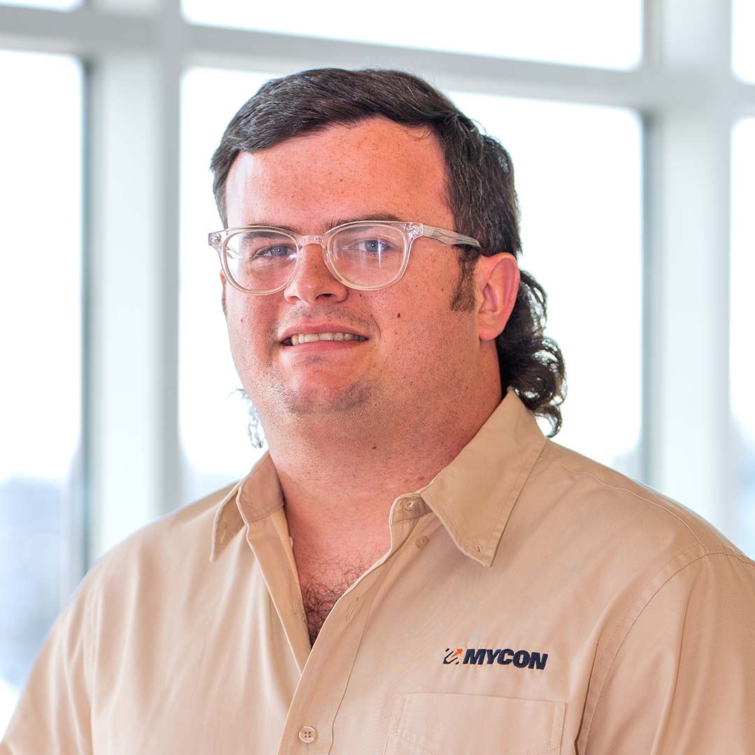A man wearing glasses and a tan MYCON shirt stands indoors in front of large windows, looking at the camera.