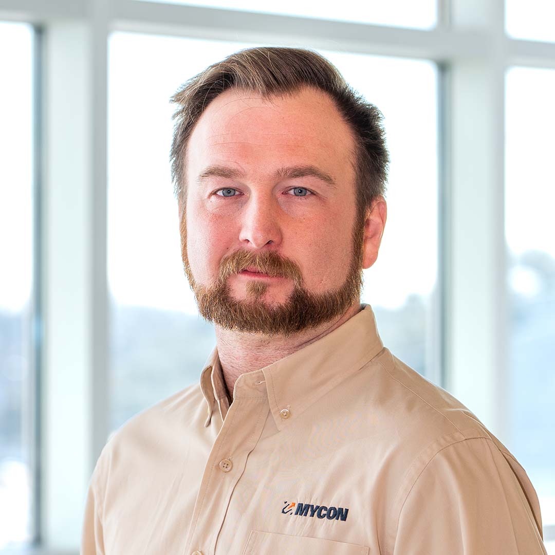 A man with light brown hair and a beard wears a tan MYCON-branded shirt, standing indoors with large windows in the background.
