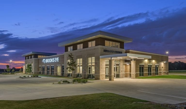 A modern, single-story medical building labeled "Brooks Eye Surgery Center" stands in an empty parking lot at dusk, with a colorful sky and nearby self storage units in the background.