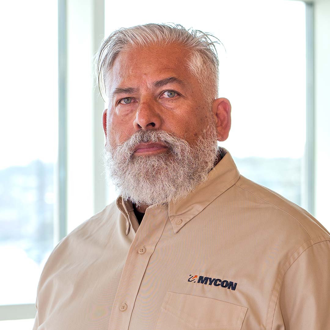 A man with gray hair and a beard wearing a beige MYCON-branded shirt stands indoors in front of large windows.