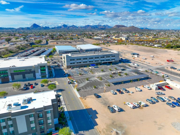 Aerial view of a commercial area featuring a multi-story office building, self storage facility, parking lots, nearby roads, and mountains visible in the background under a partly cloudy sky.