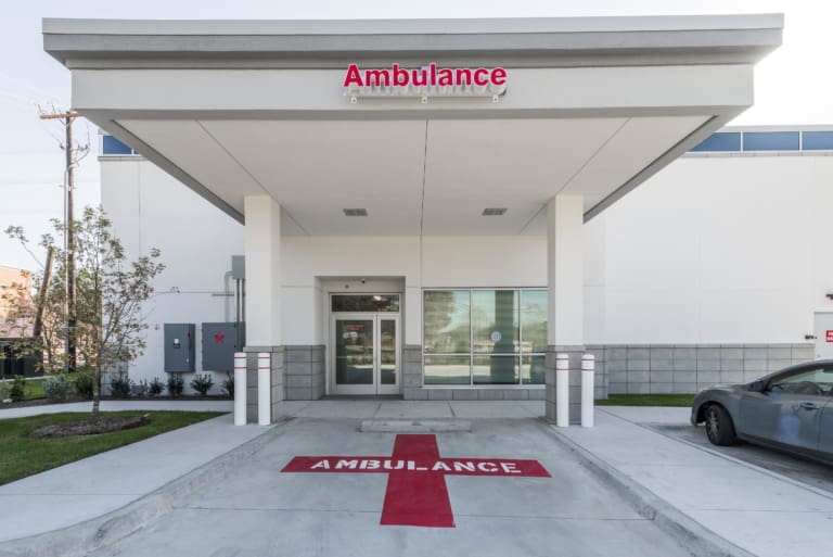 Ambulance entrance at a hospital with a red cross and the word "AMBULANCE" painted on the ground under a covered driveway, located next to a self storage facility.